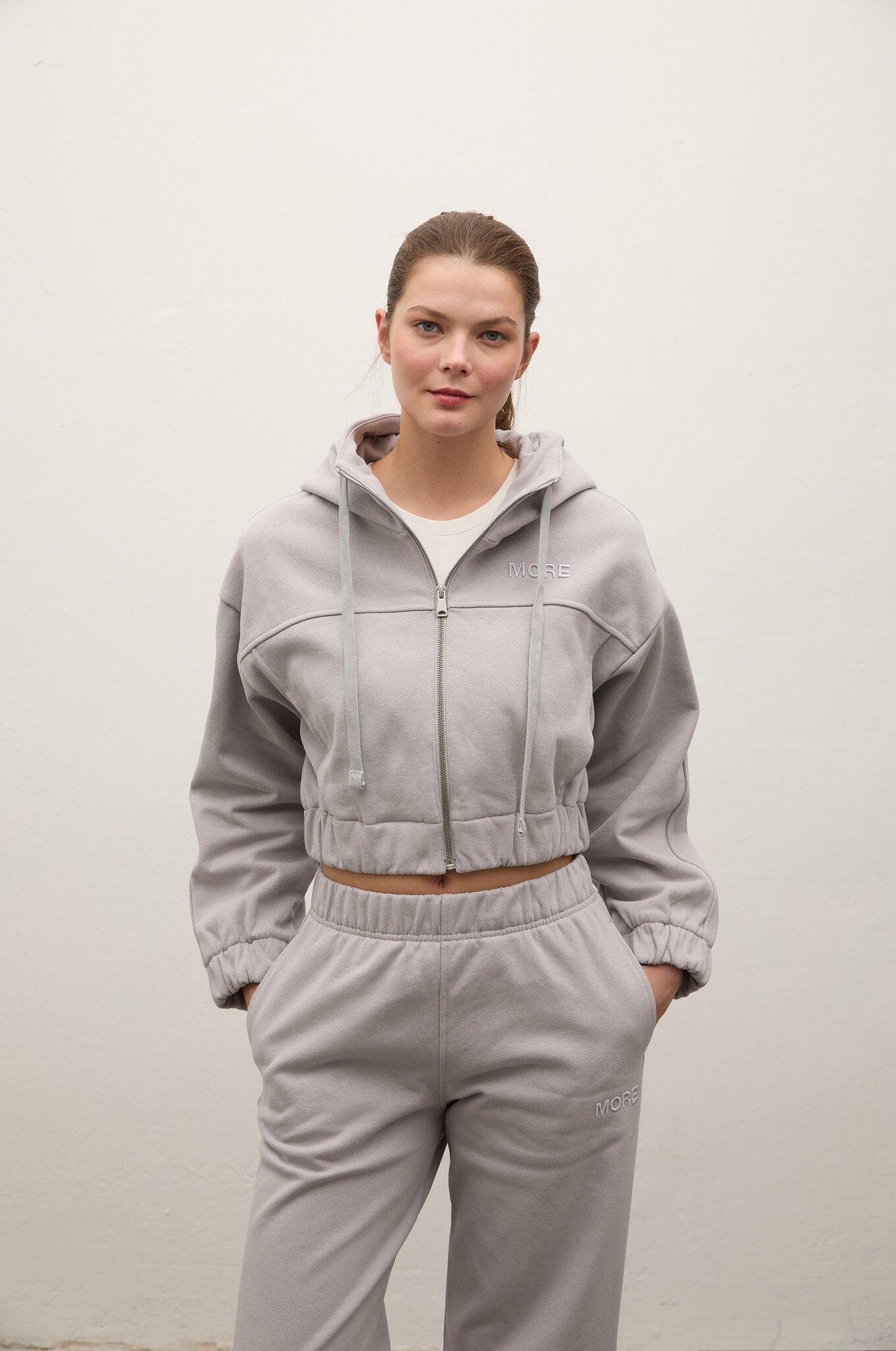 A woman modeling a light gray cropped zip-up hoodie and matching sweatpants set against a plain off-white wall.