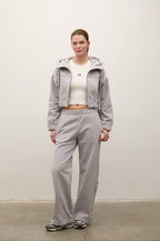 A model stands against a plain off-white wall wearing a light gray hooded jacket, matching wide-leg sweatpants, a white cropped tank top, and gray sneakers.