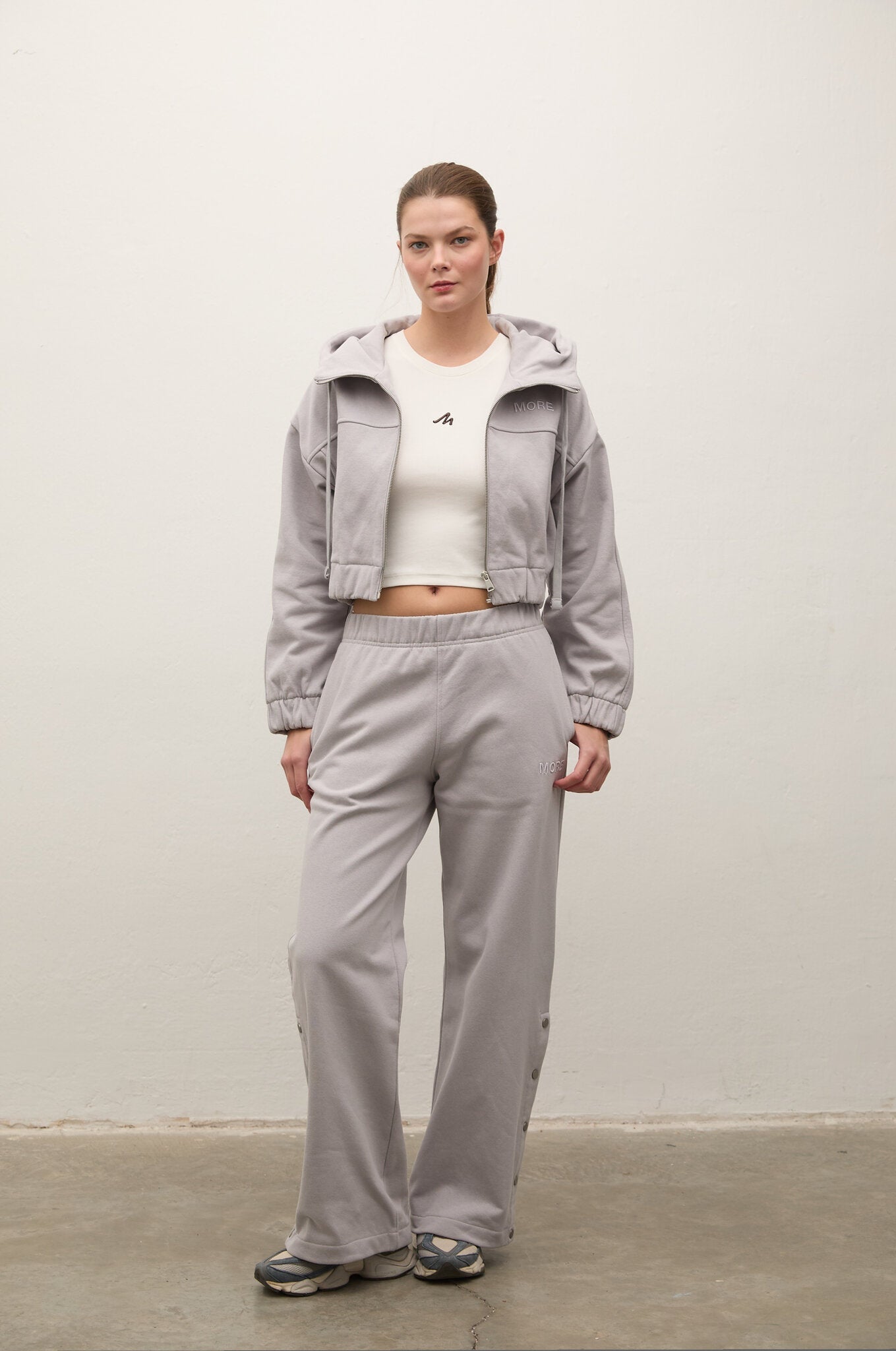 A model stands against a plain off-white wall wearing a light gray hooded jacket, matching wide-leg sweatpants, a white cropped tank top, and gray sneakers.