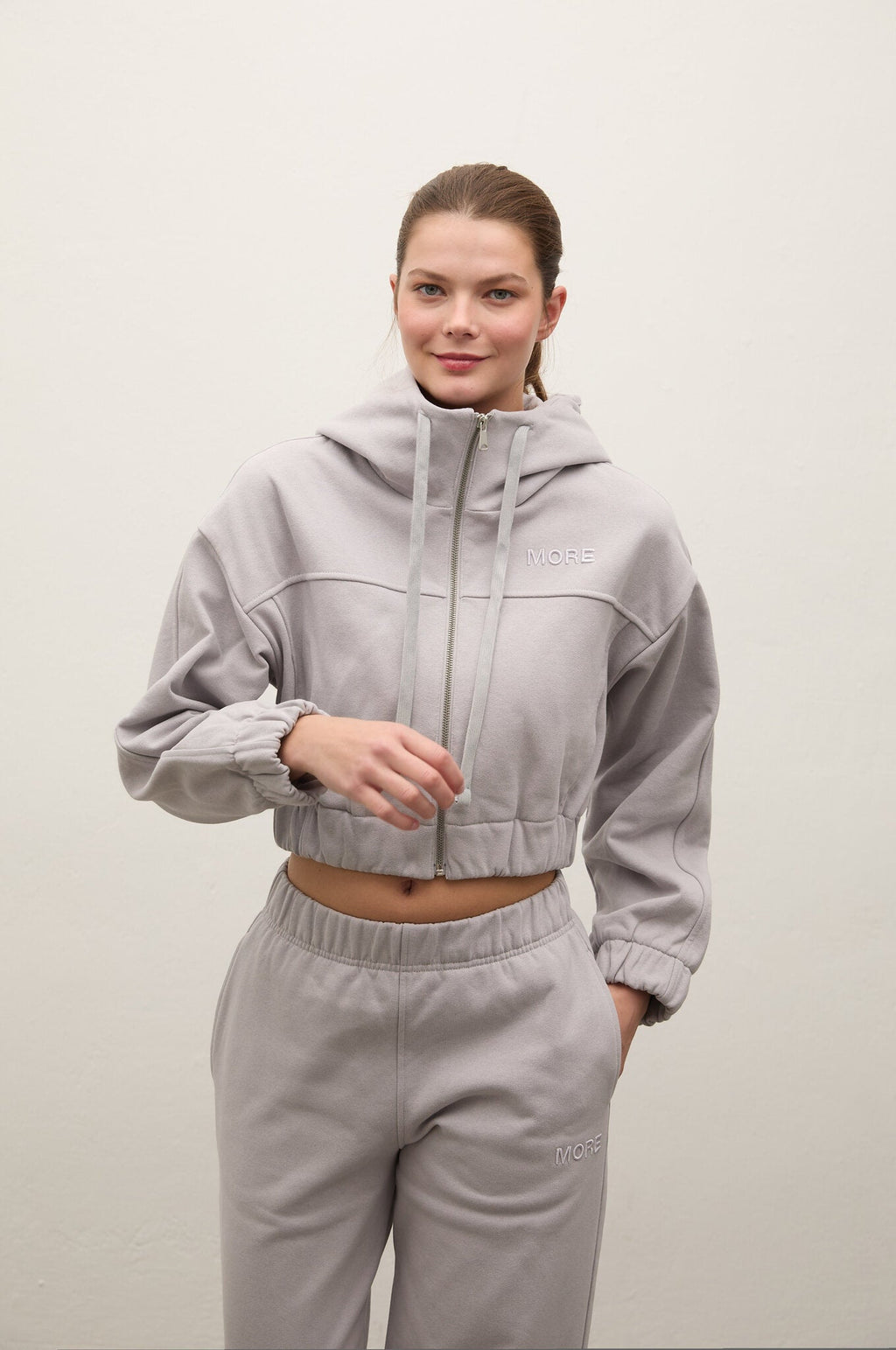 A woman modeling a light gray cropped zip-up hoodie and matching jogger sweatpants against a plain off-white background.