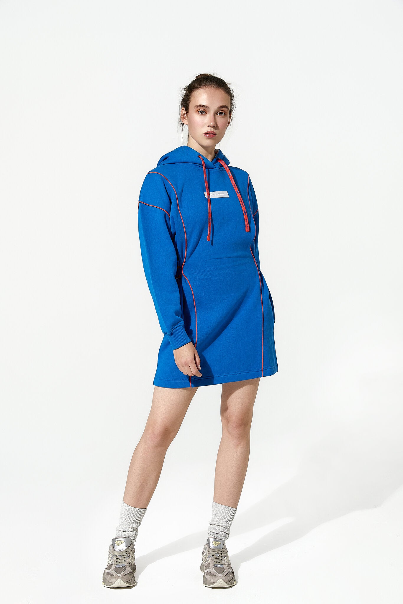 A model wearing a bright blue hooded sweatshirt dress with red drawstrings and red piping details, styled with gray socks and gray sneakers, against a plain white studio background.