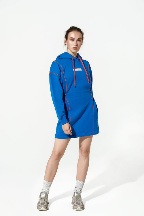 A model wearing a bright blue hooded sweatshirt dress with red drawstrings and red piping details, styled with gray socks and gray sneakers, against a plain white studio background.