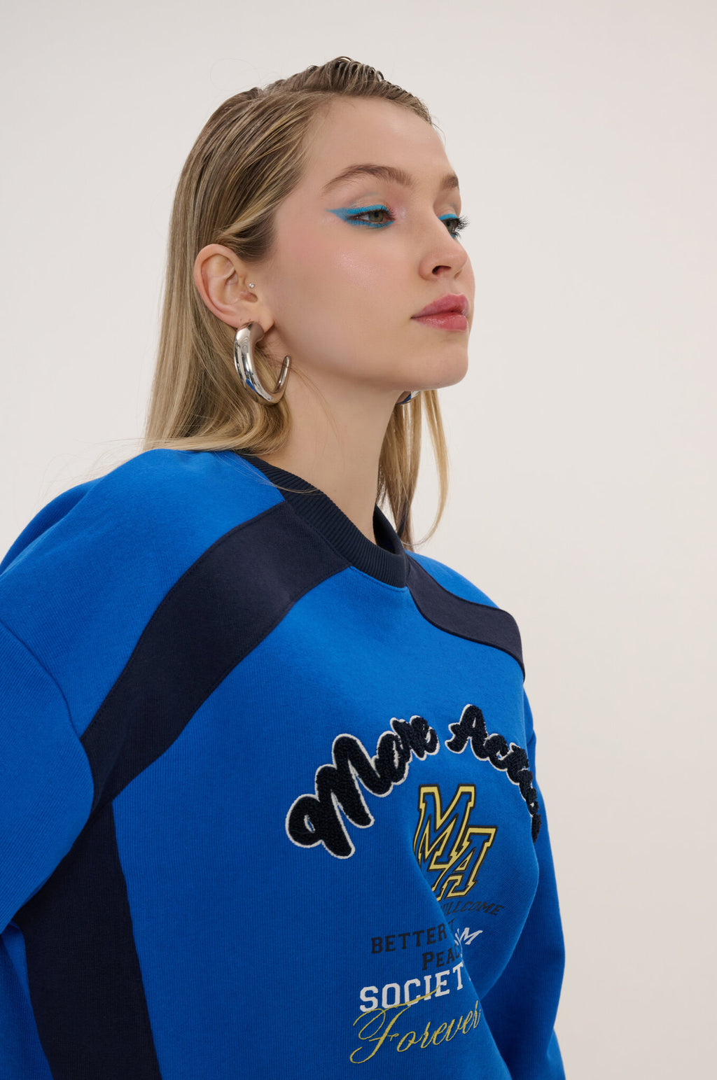 A woman wearing a bright blue and navy varsity-style sweatshirt with 'Mia Rose' and 'SOCIETY Forever' text on the front, photographed against a plain light background.