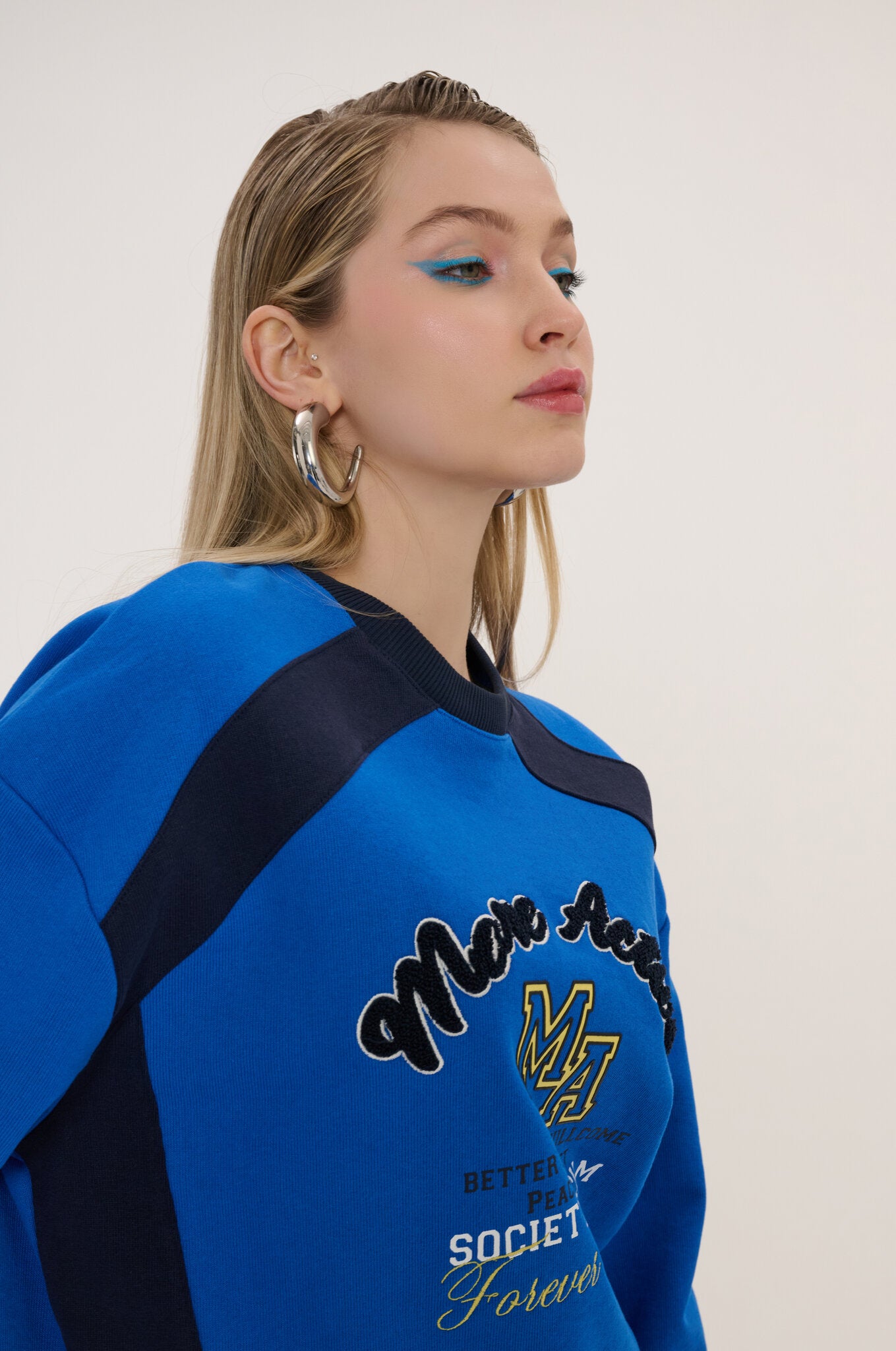 A woman wearing a bright blue and navy varsity-style sweatshirt with 'Mia Rose' and 'SOCIETY Forever' text on the front, photographed against a plain light background.