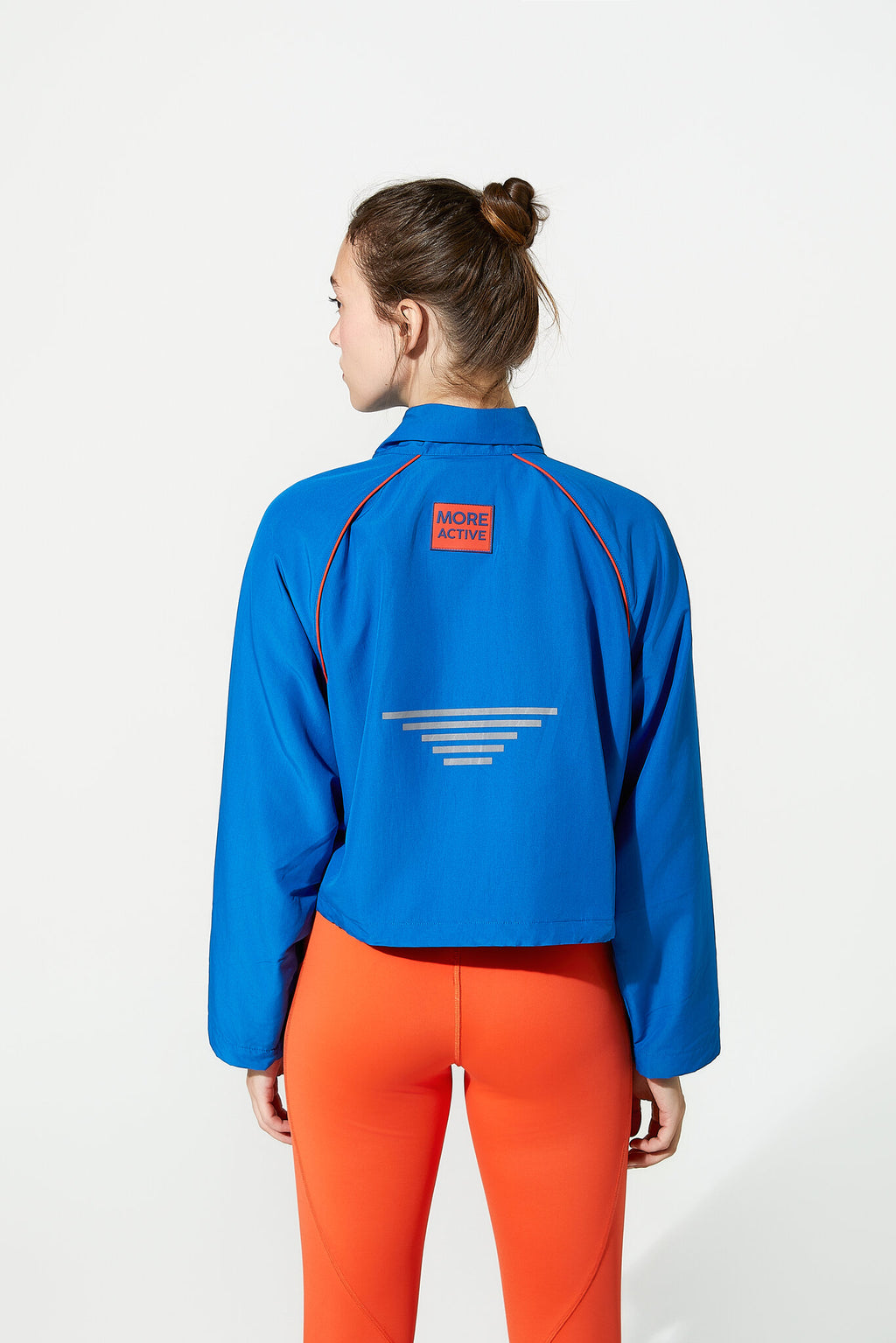 A woman shown from the back wearing a bright blue cropped athletic jacket with a small orange 'MORE ACTION' patch on the upper back and a white geometric stripe graphic below it, paired with bright orange leggings against a clean white studio background.
