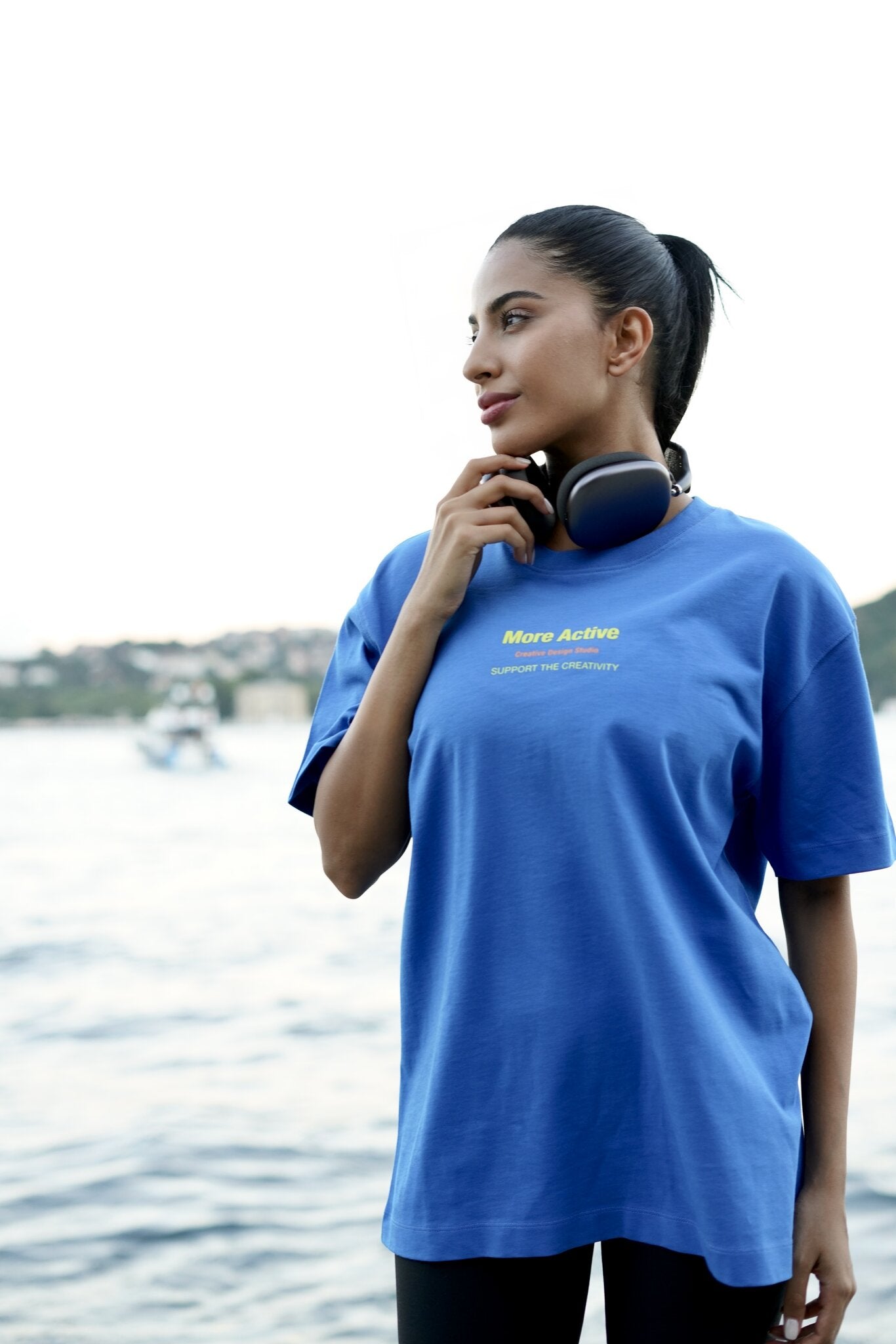 A woman wearing an oversized blue t-shirt with yellow 'More Active' text, standing outdoors near the water with headphones around her neck.