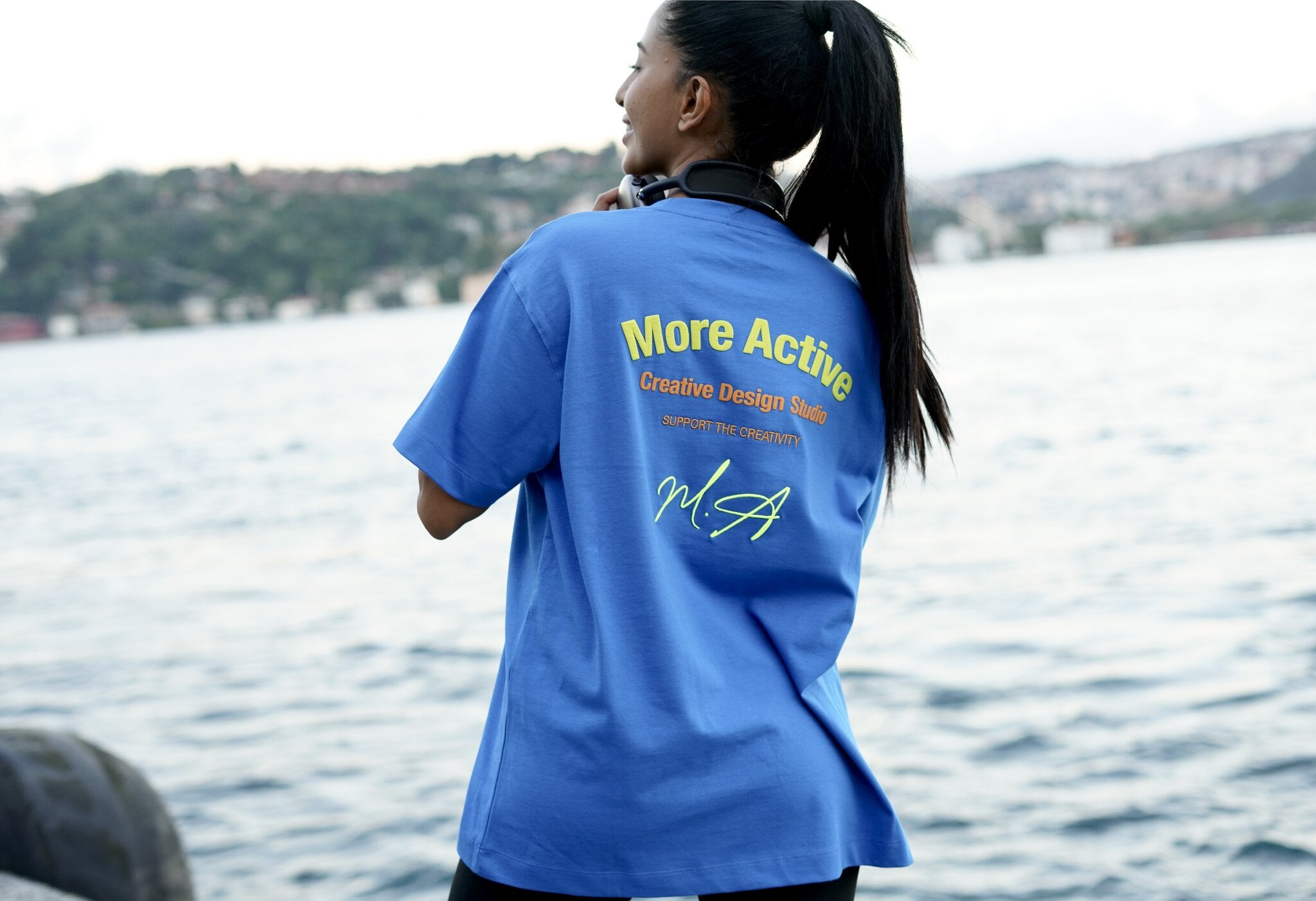 A woman stands with her back to the camera wearing a bright blue t-shirt with yellow and white text on the back that reads 'More Active Creative Design Team'. The background shows a body of water and distant hills.