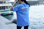 A woman stands on a waterfront dock wearing a bright blue t-shirt with yellow and white text on the back that reads 'More Active Creative Design Studio'.