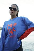 A woman wearing oversized sunglasses and a blue long-sleeve sweatshirt with red graphic lettering and red underlayer sleeves, photographed outdoors near water.