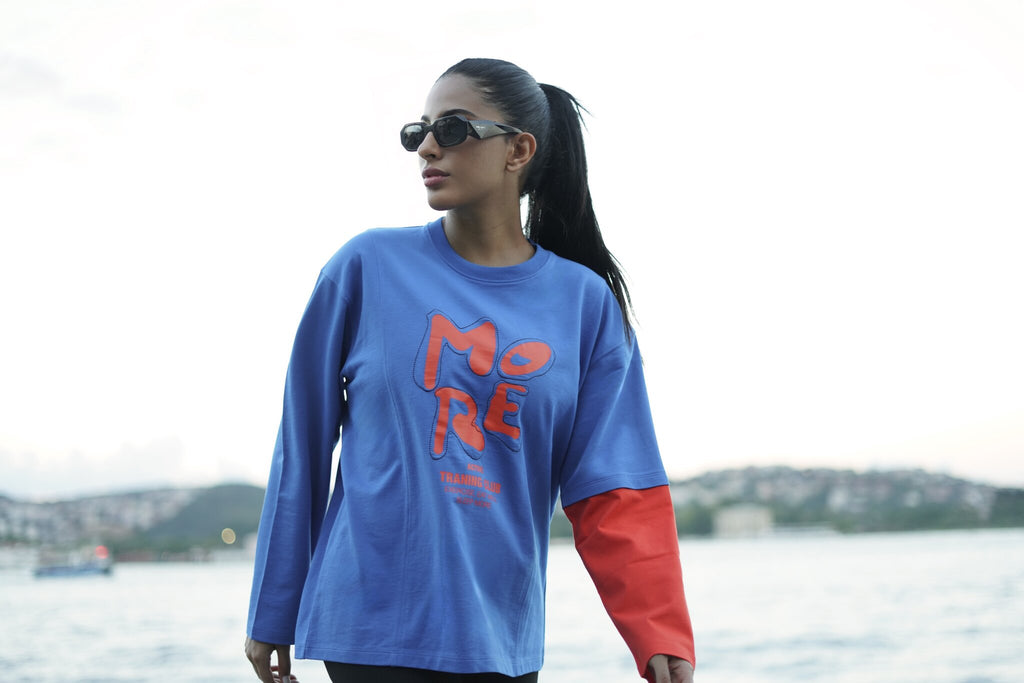 A woman wearing a blue long-sleeve graphic t-shirt with red and orange lettering and a red sleeve, standing outdoors near a body of water with a hazy sky and distant hills in the background.