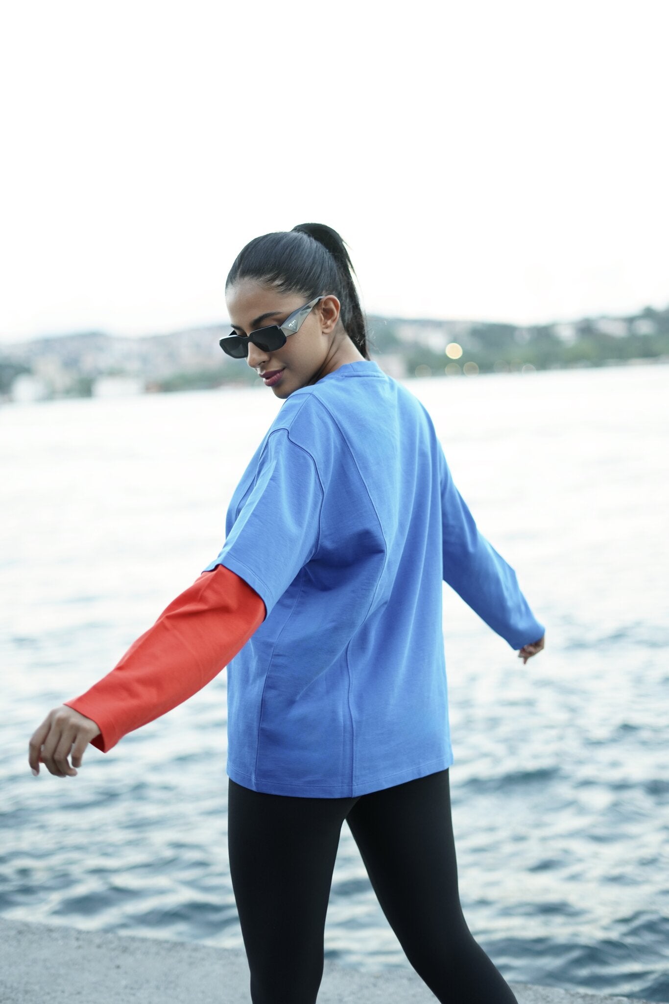 A woman wearing a light blue long-sleeve athletic top with contrasting red sleeves and black leggings, standing by the water with a blurred background.
