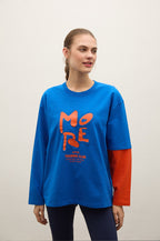 A woman modeling a long-sleeve graphic t-shirt with a vibrant blue body, a red sleeve, and a large red 'MORE' graphic on the front, set against a plain light gray background.