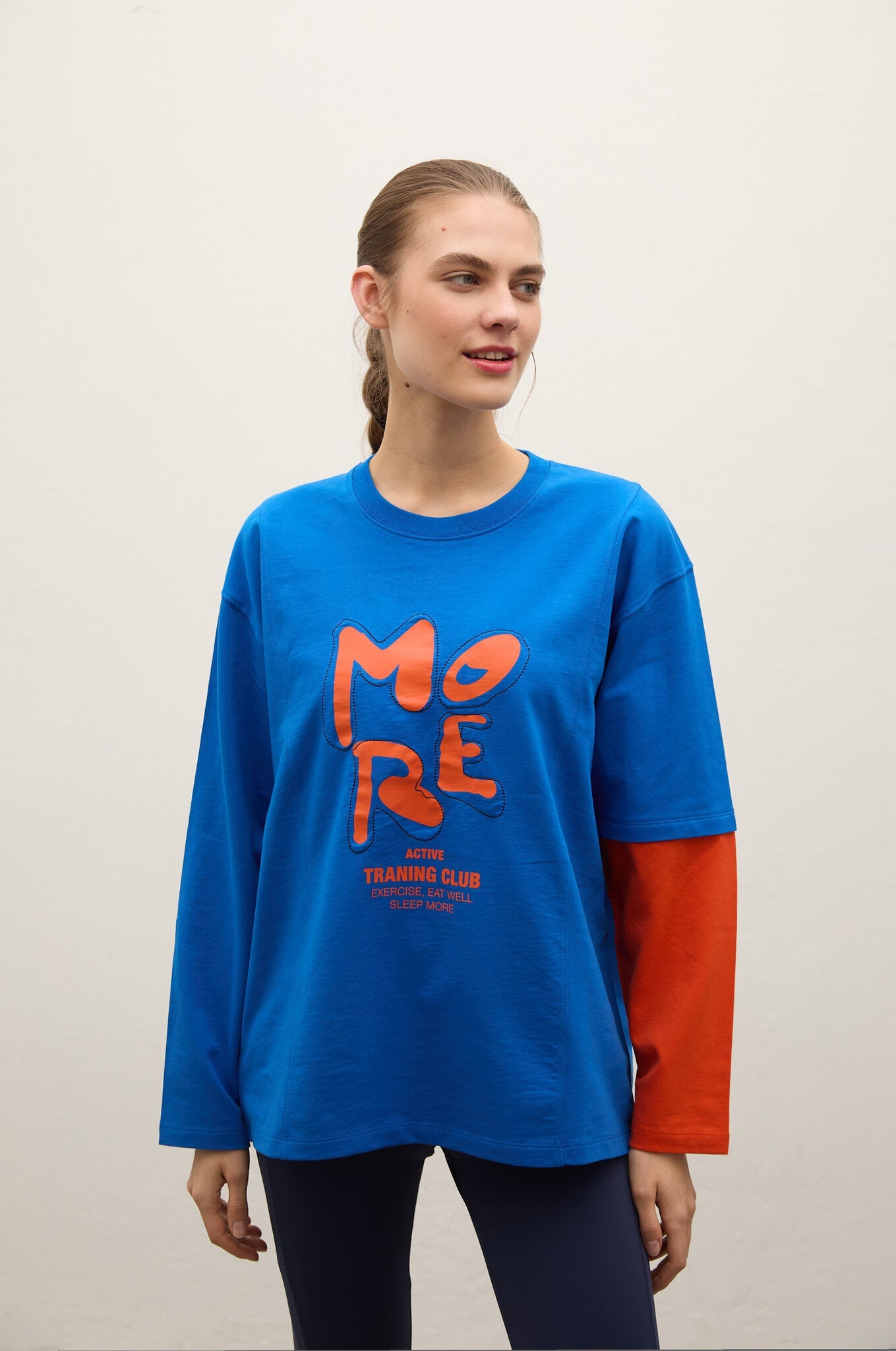 A woman modeling a long-sleeve graphic t-shirt with a vibrant blue body, a red sleeve, and a large red 'MORE' graphic on the front, set against a plain light gray background.