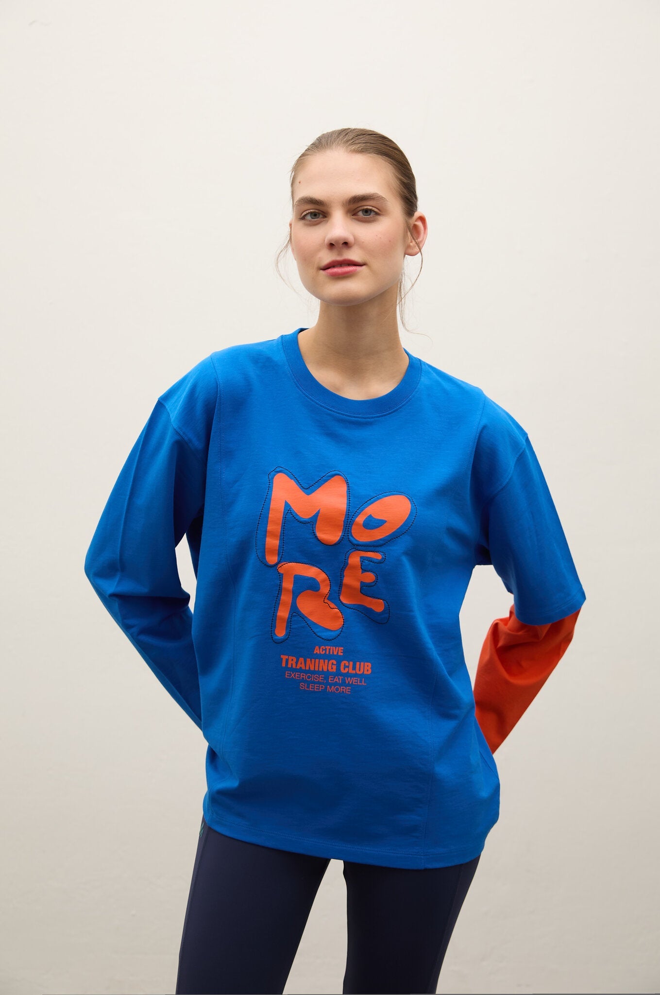 A woman modeling a bright blue long-sleeve t-shirt with a large orange graphic on the front that reads 'MORE TRAINING CLUB'. The background is a clean, neutral off-white studio wall.