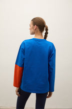 A woman viewed from behind wearing a long-sleeve colorblock shirt with a vibrant blue body and orange sleeves, paired with dark pants, against a plain off-white background.