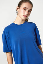 A woman wearing a bright blue short-sleeve crewneck t-shirt with the word 'MORE' printed in a matching blue font on the left chest, set against a plain light gray background.