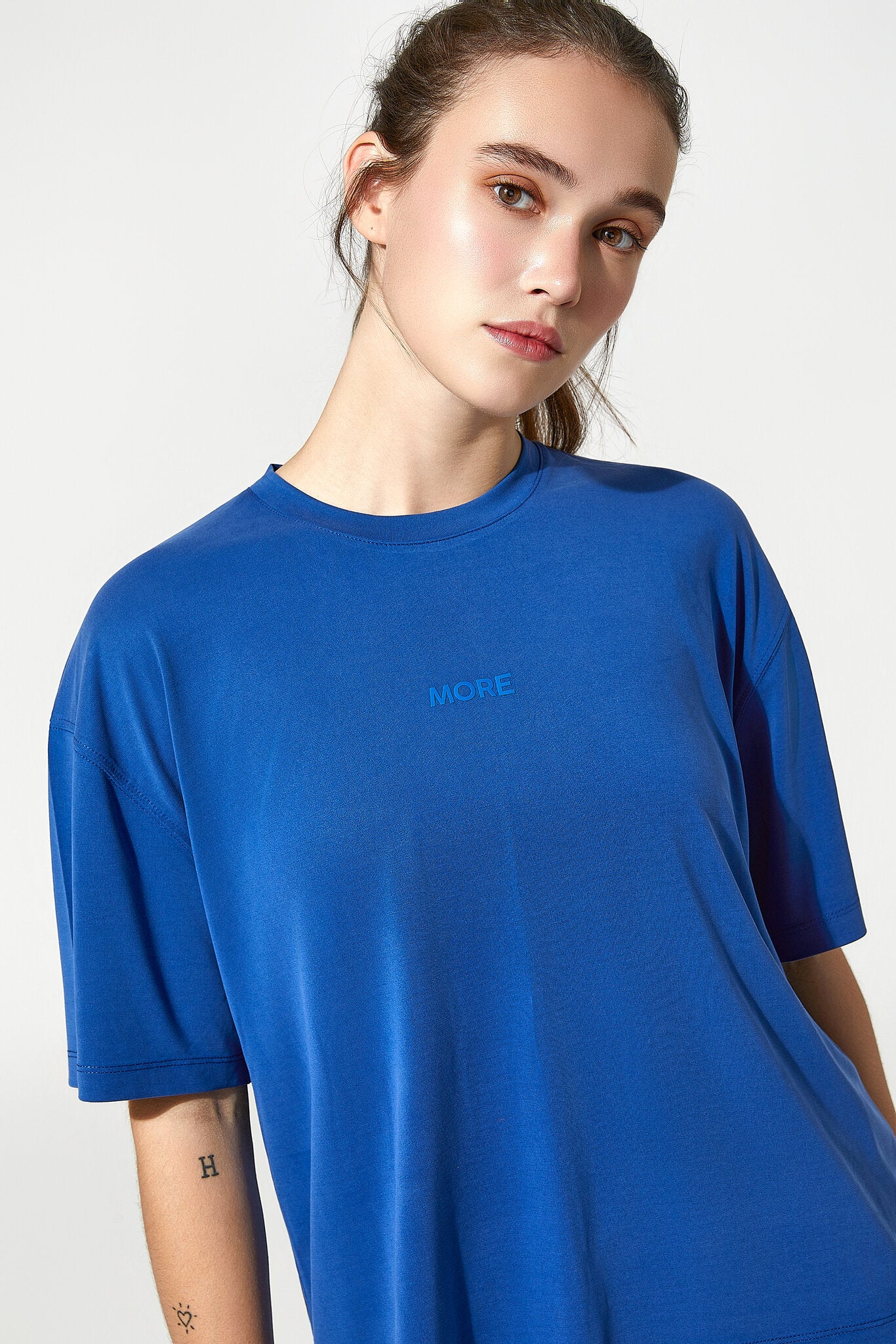 A woman wearing a bright blue short-sleeve crewneck t-shirt with the word 'MORE' printed in a matching blue font on the left chest, set against a plain light gray background.