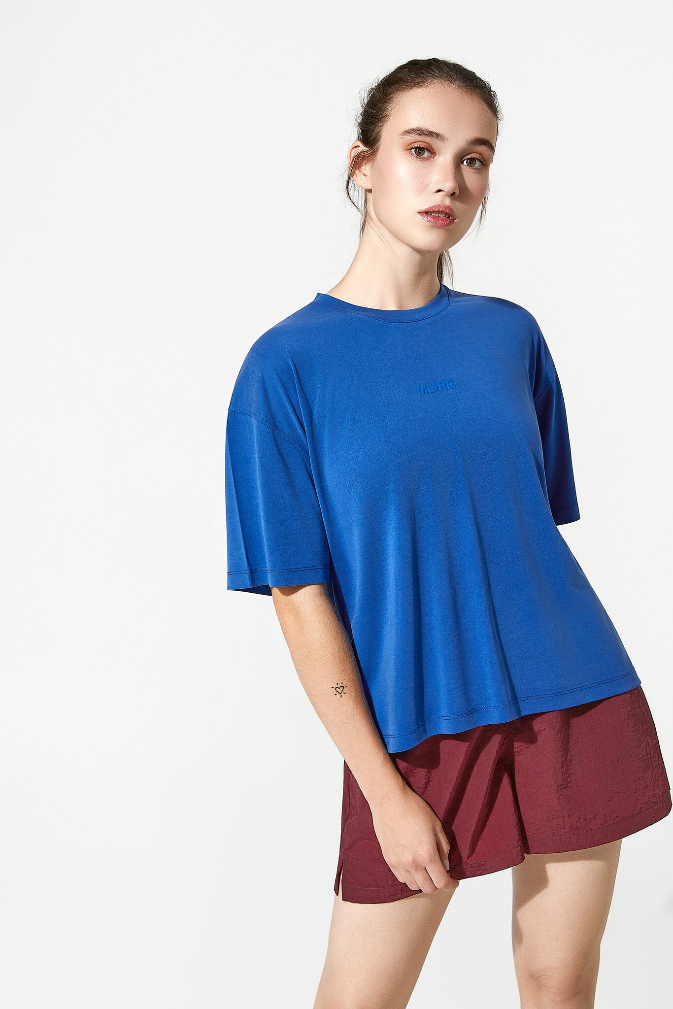 A woman modeling a vibrant blue short-sleeve crewneck t-shirt with a subtle tonal logo on the chest, paired with burgundy shorts, against a clean white studio background.