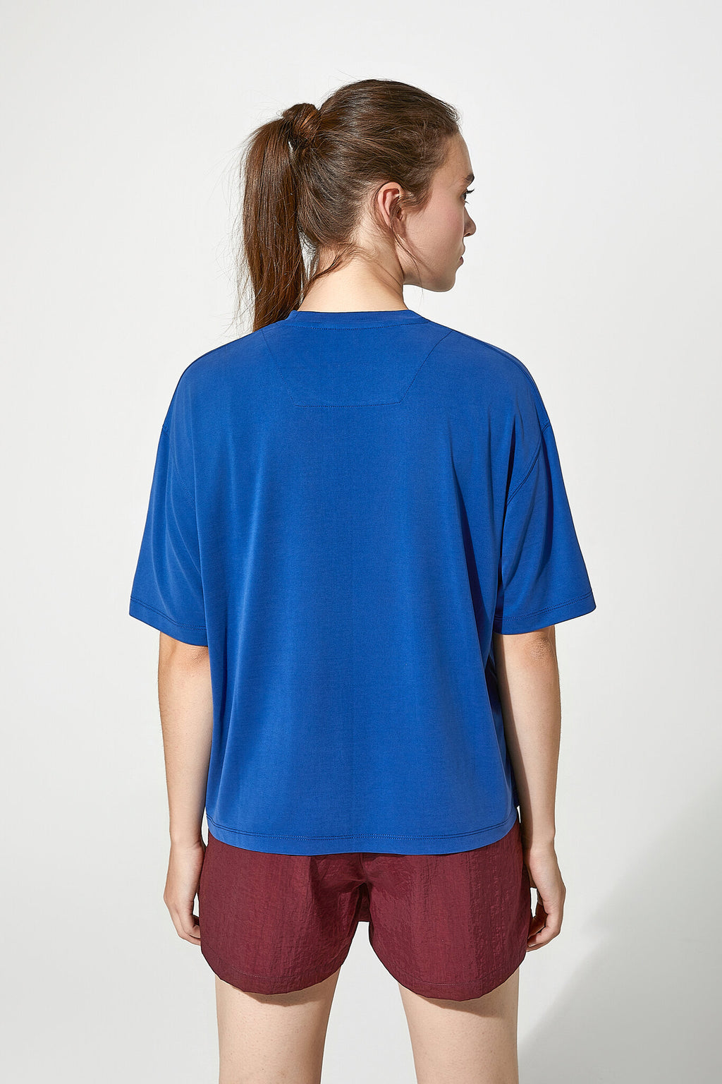 A woman shown from the back wearing a loose-fitting royal blue short-sleeve t-shirt and burgundy shorts against a plain light gray studio background.
