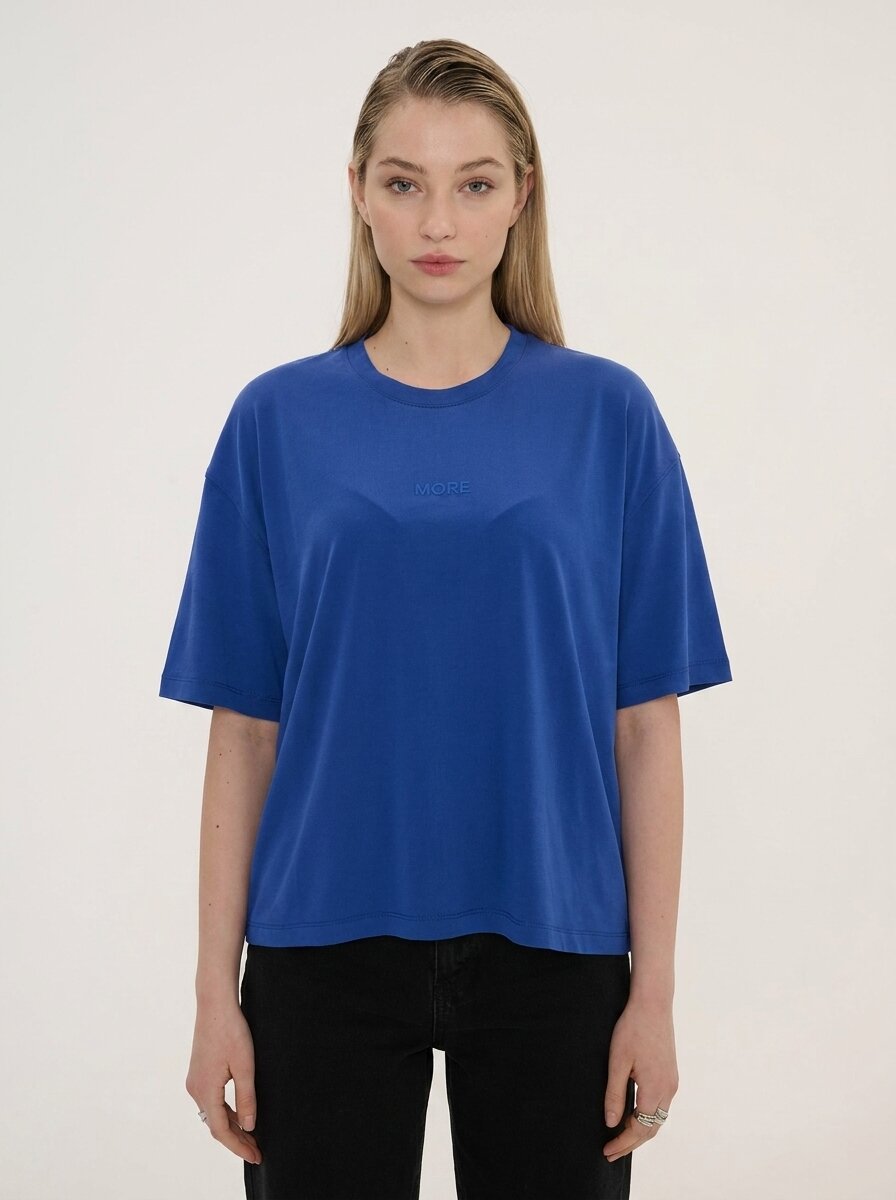 A woman modeling an oversized royal blue short-sleeve t-shirt with a subtle tonal embroidered logo on the chest, paired with black pants, against a plain off-white background.