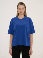 A woman modeling an oversized royal blue short-sleeve t-shirt with a subtle tonal embroidered logo on the chest, paired with black pants, against a plain off-white background.