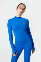 A woman modeling a bright blue long-sleeve mock-neck athletic top and matching leggings against a light gray studio background. A small red circular logo is visible on the left chest.