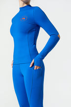 A woman wearing a bright blue long-sleeve athletic top and matching leggings against a plain light gray studio background. A small orange circular logo is visible on the left chest.