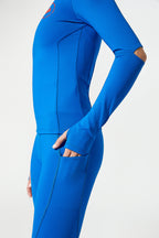 A close-up side view of a person wearing a bright blue full-body athletic suit or wetsuit with long sleeves, fitted legs, and a small orange logo near the upper chest, against a clean light gray studio background.