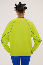 A person shown from the back wearing a bright neon yellow-green crewneck sweatshirt with contrasting blue piping details, paired with blue leggings, against a plain light background.
