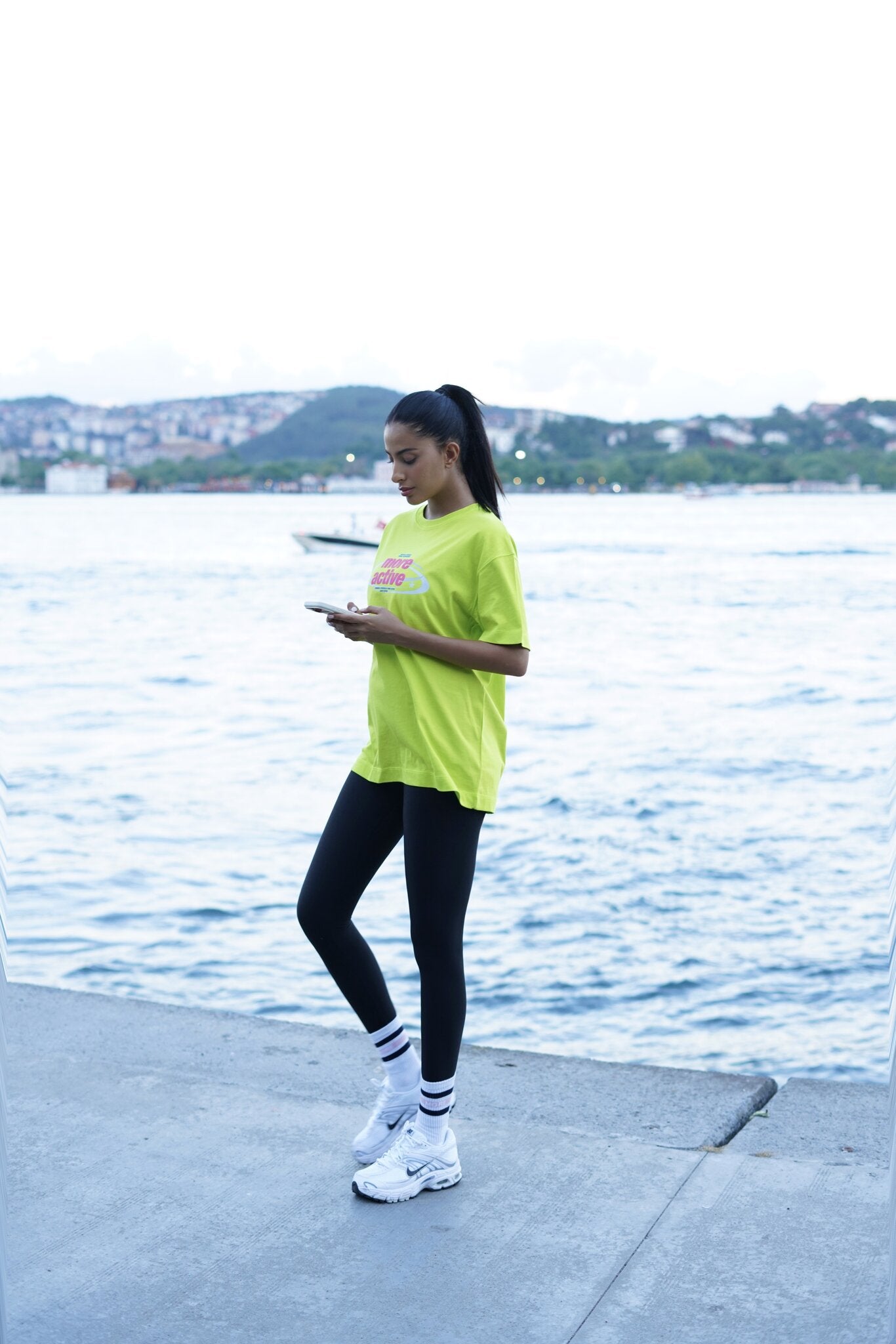 A woman stands on a concrete pier by the water, wearing a bright neon yellow-green oversized T-shirt, black leggings, white socks, and white sneakers, while looking down at a phone.