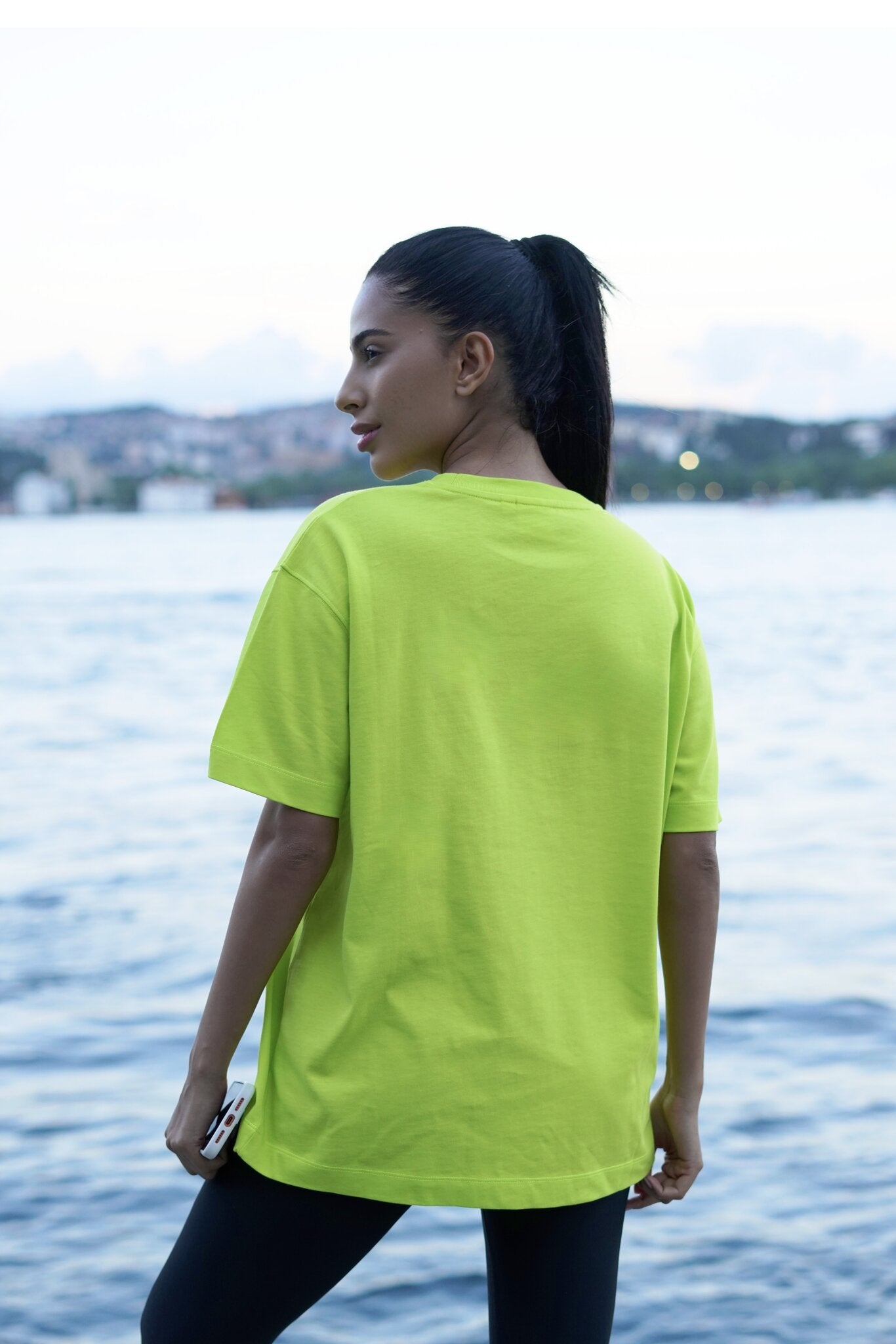 A woman with her hair in a ponytail stands by the water wearing an oversized neon yellow-green t-shirt and black leggings, viewed from behind.