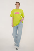 A model wearing a bright neon yellow-green oversized t-shirt with a colorful retro graphic on the chest, paired with light-wash wide-leg jeans and white sneakers, against a plain light gray studio background.