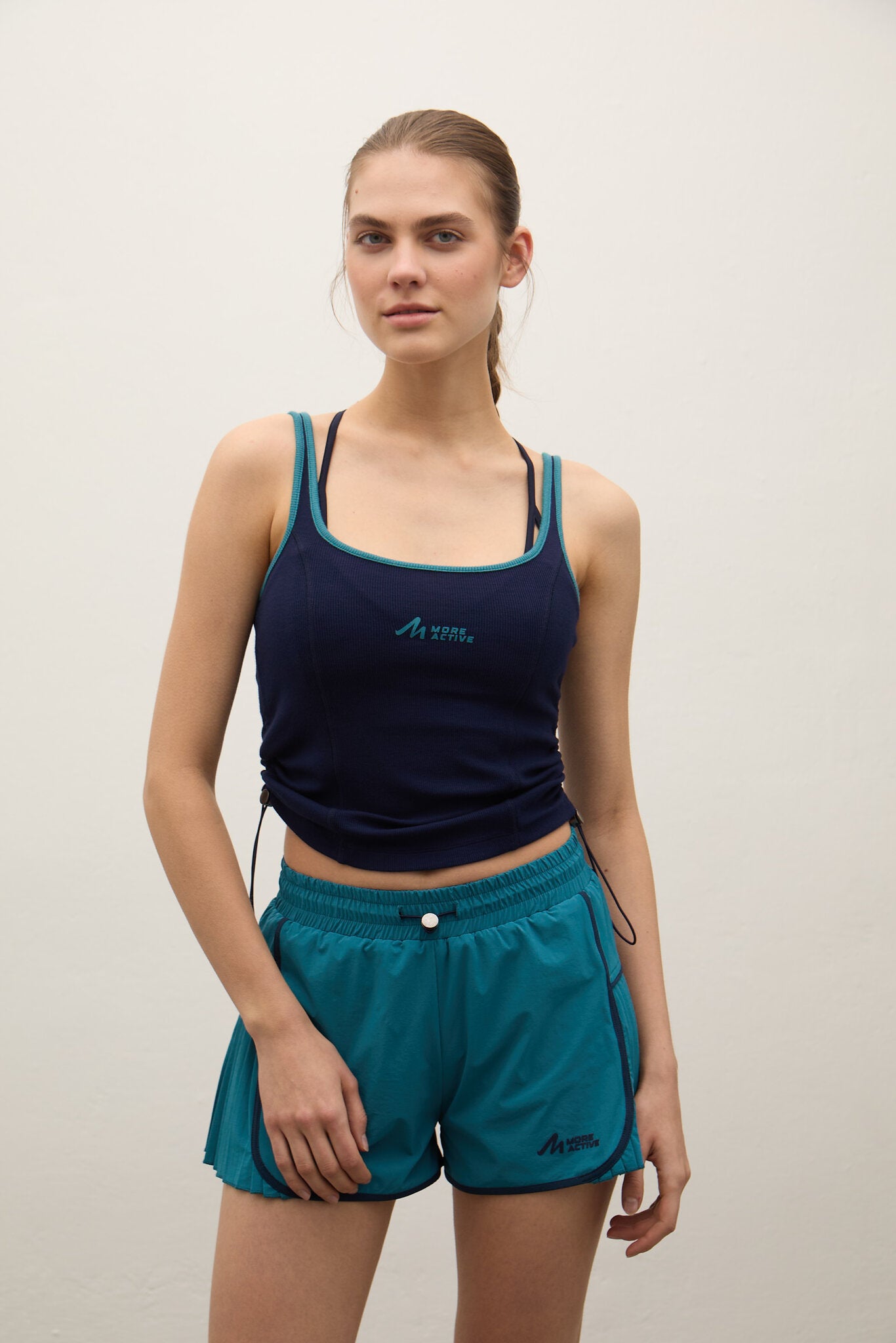 A woman modeling a navy blue athletic tank top and teal athletic shorts against a plain off-white background.