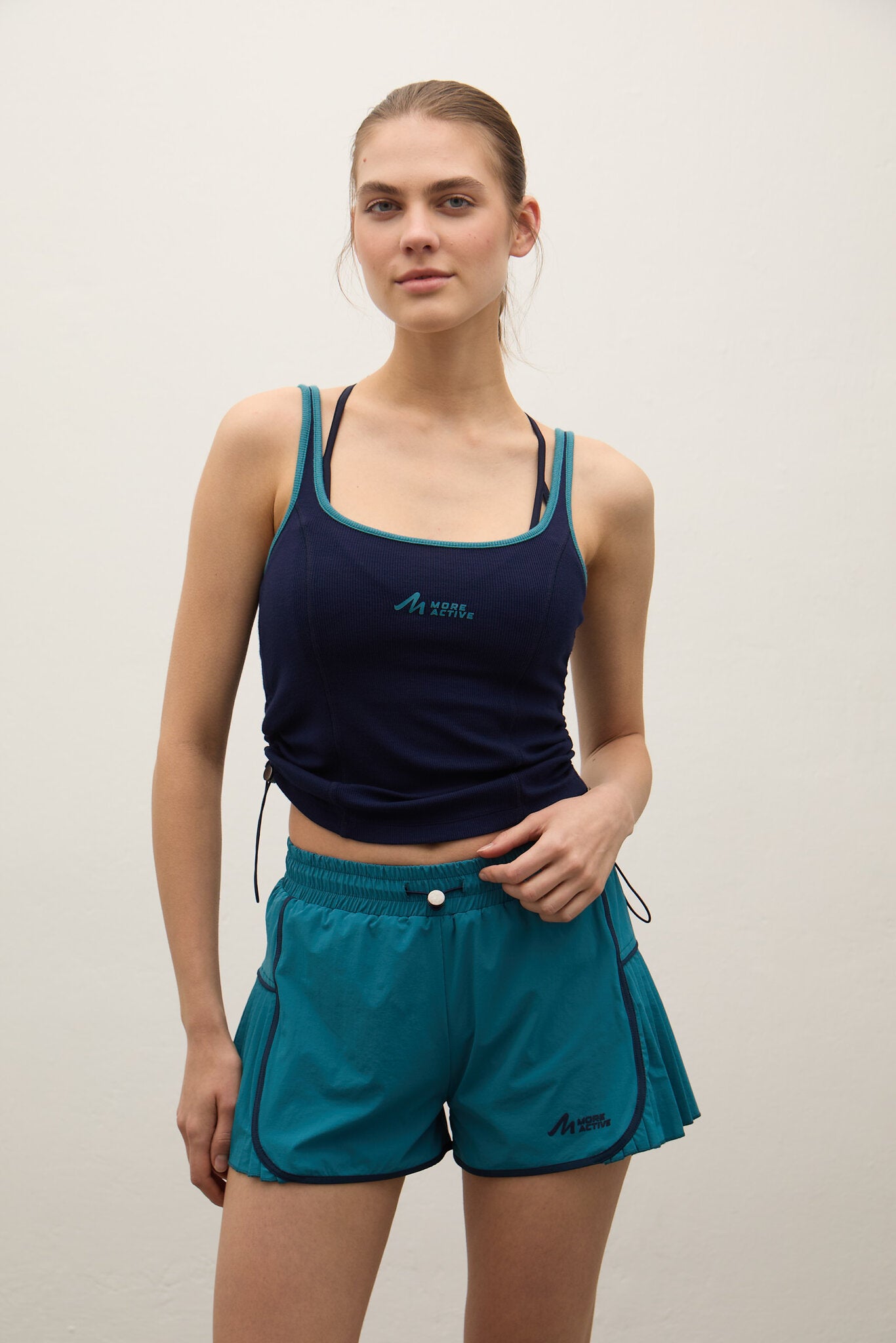 A woman modeling a navy blue athletic tank top and teal athletic shorts with teal trim and drawstrings, standing against a plain off-white wall.