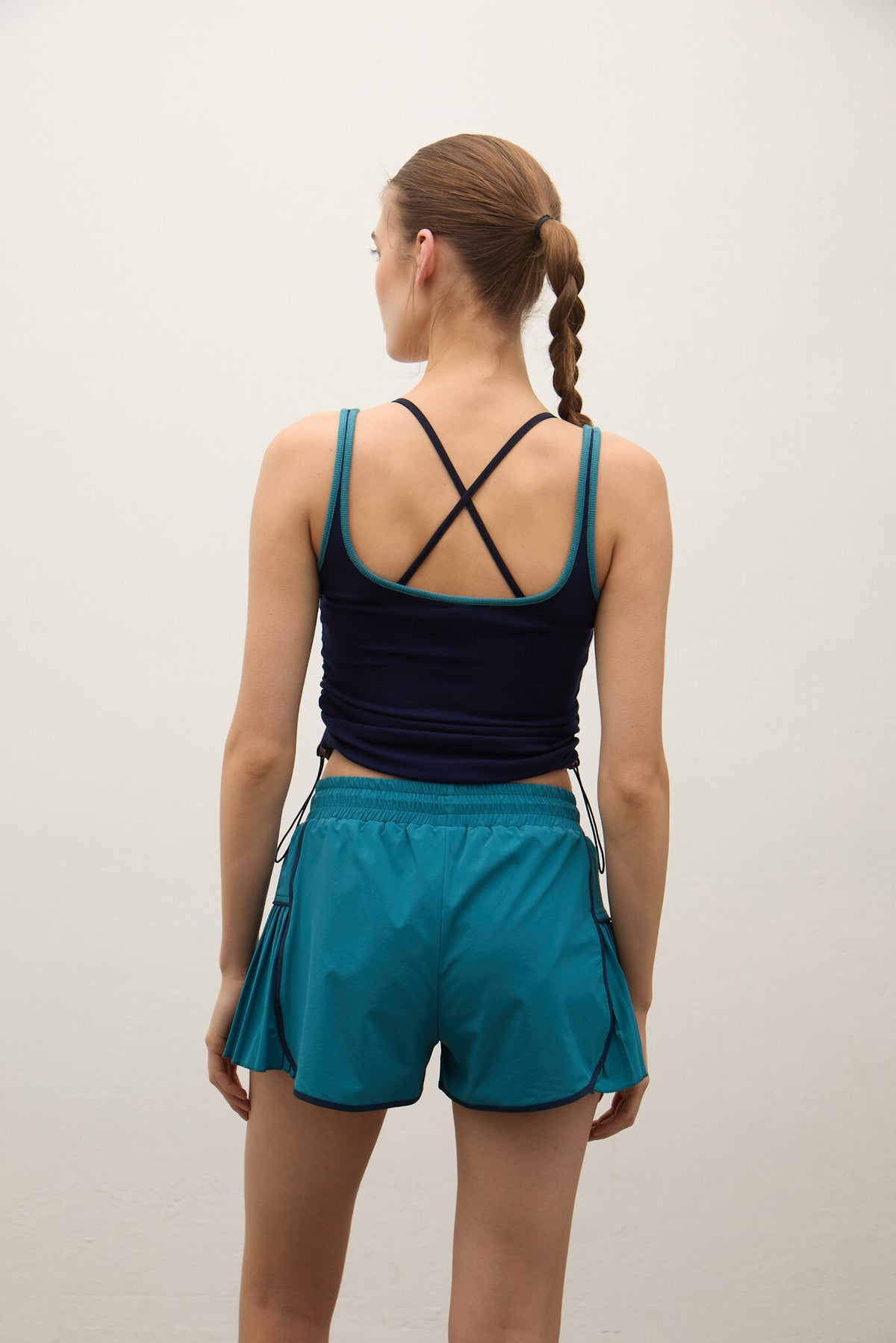 A woman shown from the back wearing a dark navy athletic tank top with crisscross straps and teal-blue athletic shorts, standing against a plain light gray wall.