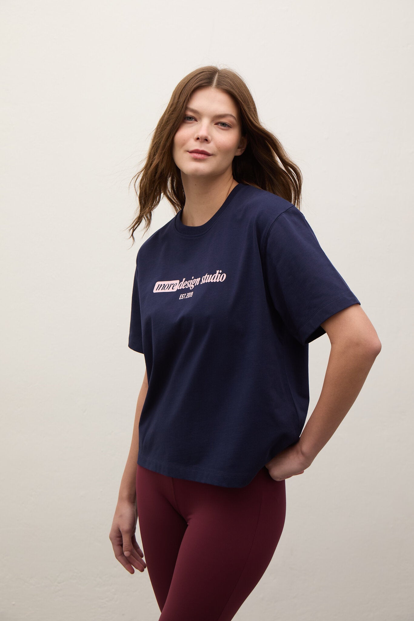 A woman modeling a navy blue short-sleeve t-shirt with a white graphic print on the chest, paired with burgundy leggings, against a plain off-white wall.