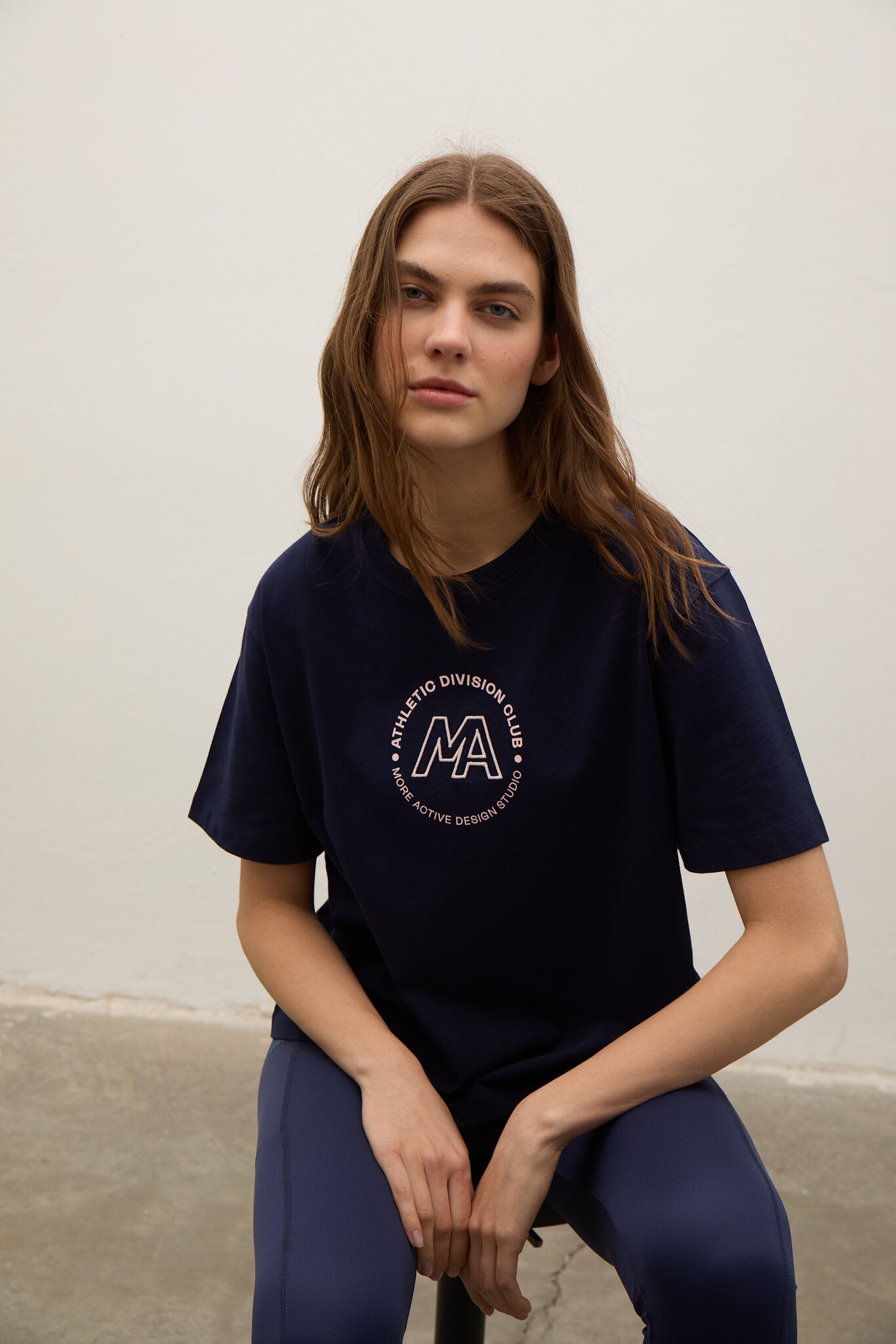 A woman with long light brown hair sits against a plain off-white wall wearing a navy blue t-shirt with a circular white graphic on the chest and matching navy blue leggings.