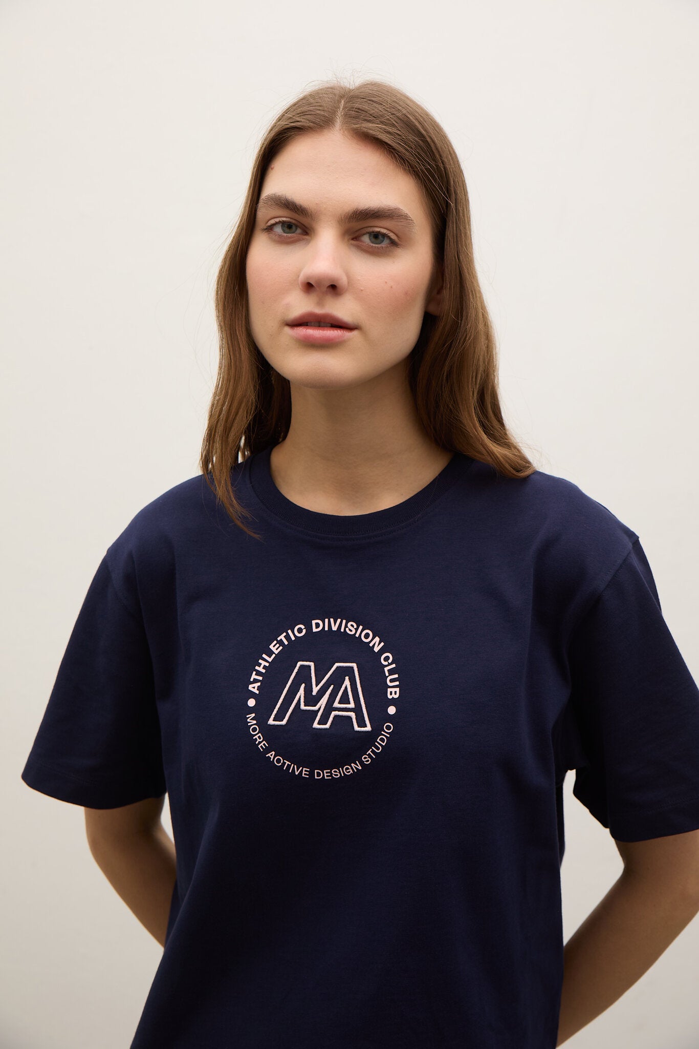 A woman with light brown hair wearing a navy blue crewneck t-shirt with a white circular graphic on the chest that reads 'ATHLETIC DIVISION CLUB' around a large 'MA' monogram.