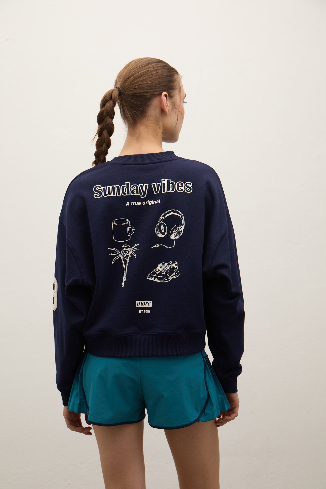 Person wearing a navy sweatshirt with 'Sunday vibes' text and graphics on the back, standing against a plain background.