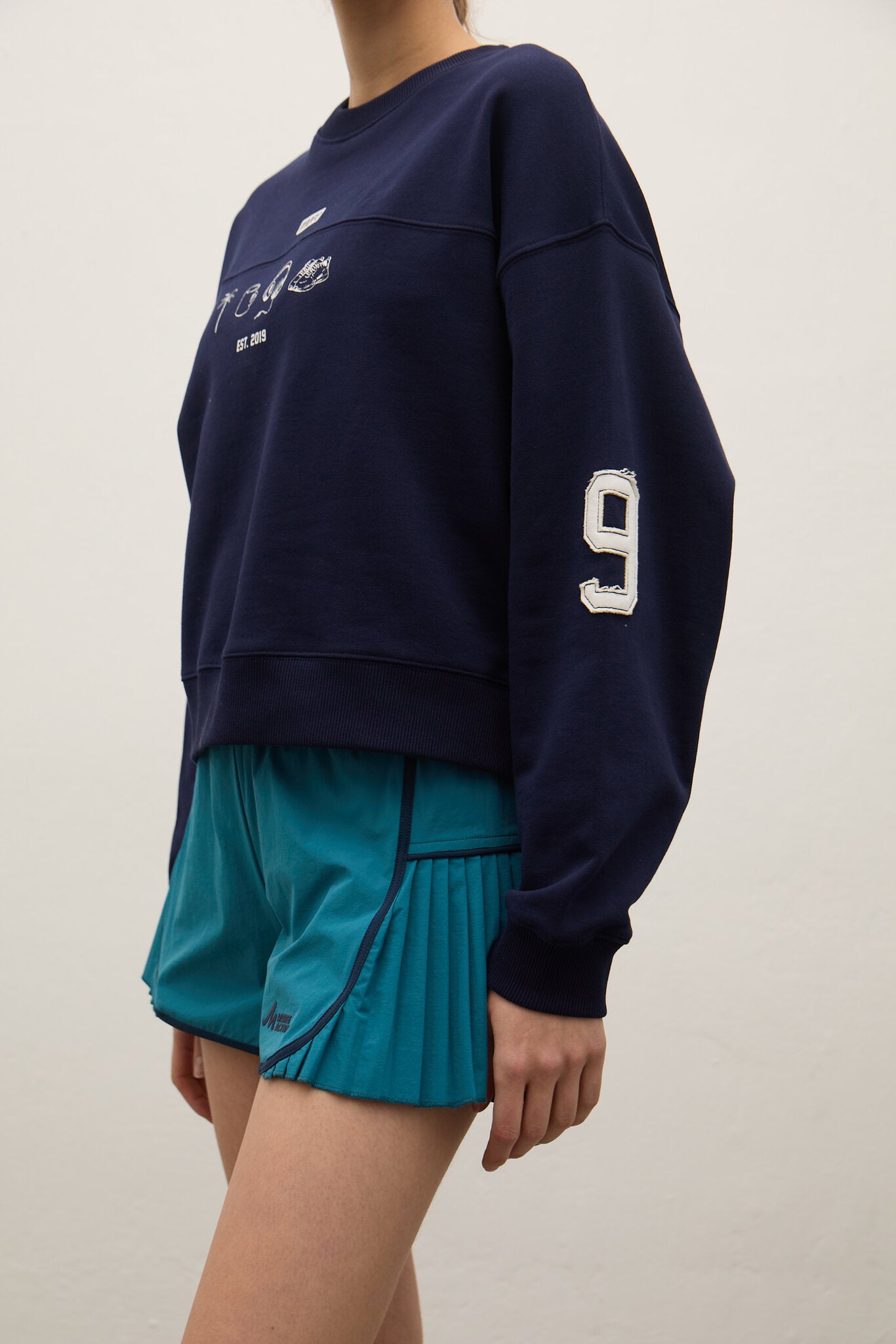Person wearing a navy blue sweatshirt with a number and teal shorts on a plain background