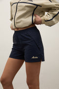 Person wearing navy shorts with a logo and a beige jacket on a plain background