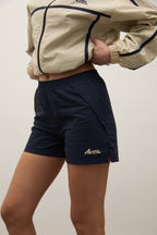 Person wearing navy shorts with a logo and a beige jacket on a plain background