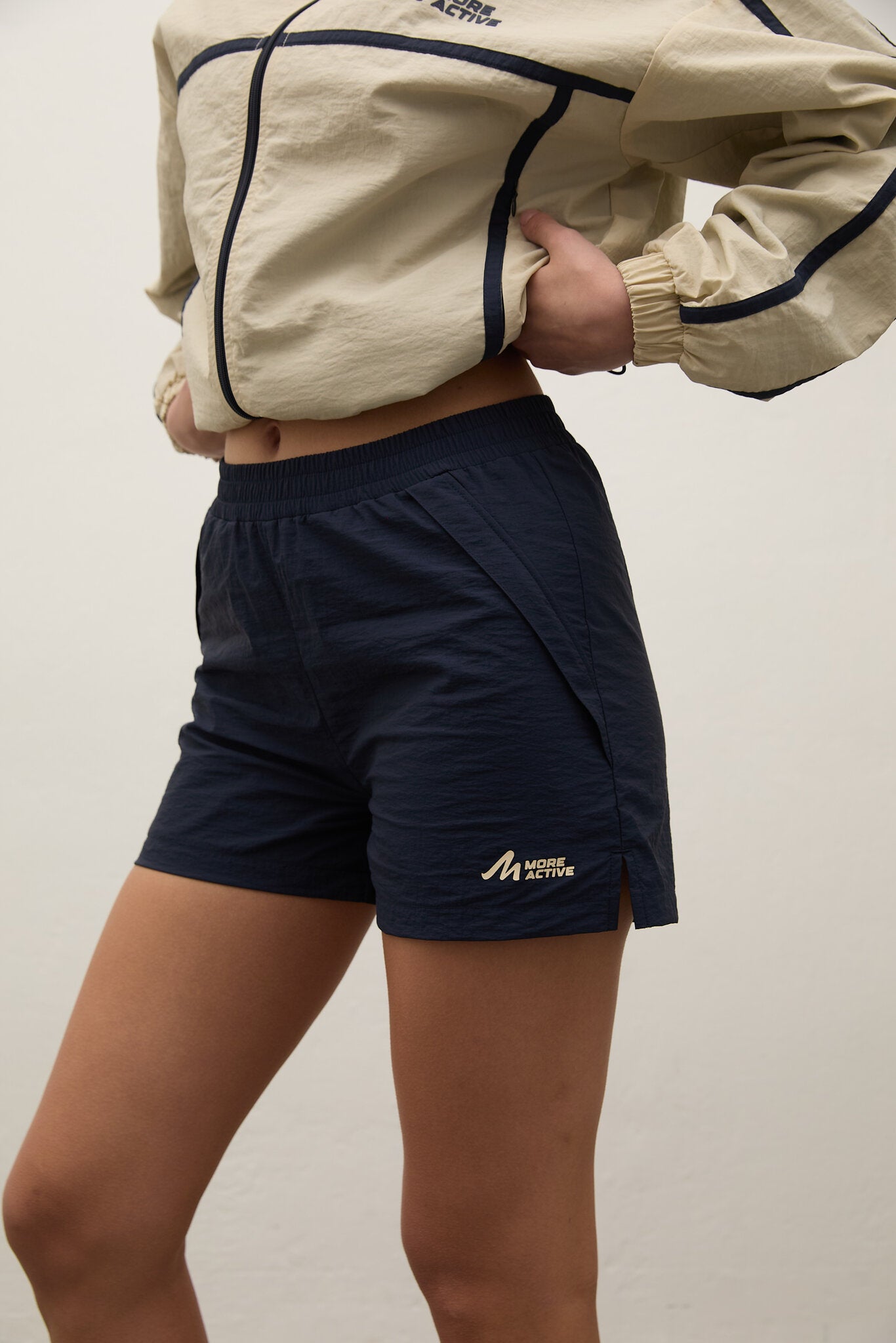 Person wearing navy shorts with a logo and a beige jacket on a plain background