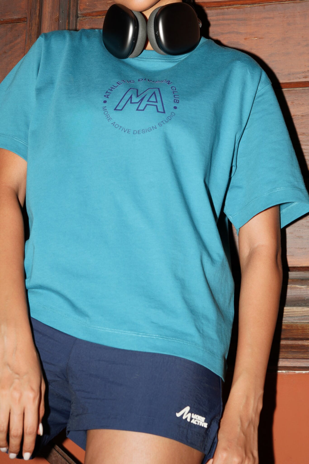 A person wearing a teal short-sleeve graphic T-shirt and navy athletic shorts, with black headphones around the neck, against a brown wooden background.