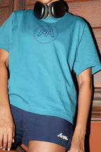 A person wearing a teal short-sleeve graphic T-shirt and navy athletic shorts, with black headphones around the neck, against a brown wooden background.