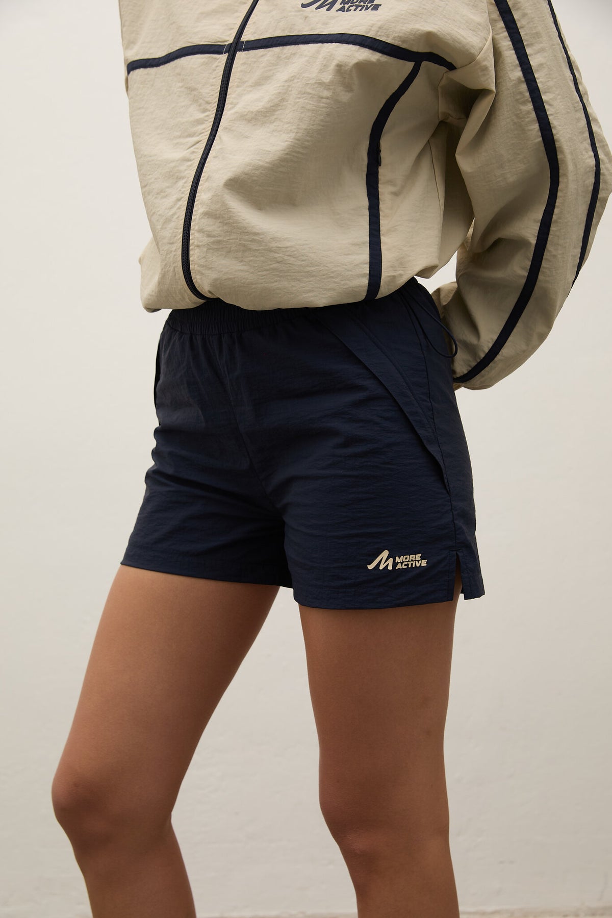 Person wearing navy shorts with a beige jacket, standing against a plain background