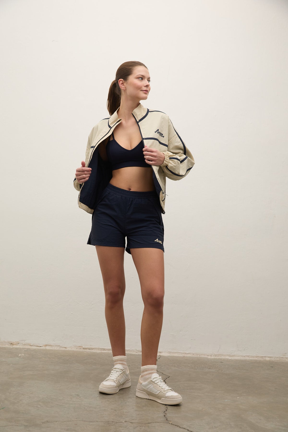 A woman modeling a beige and navy athletic jacket, navy shorts, a navy sports bra, and white sneakers against a plain off-white wall.