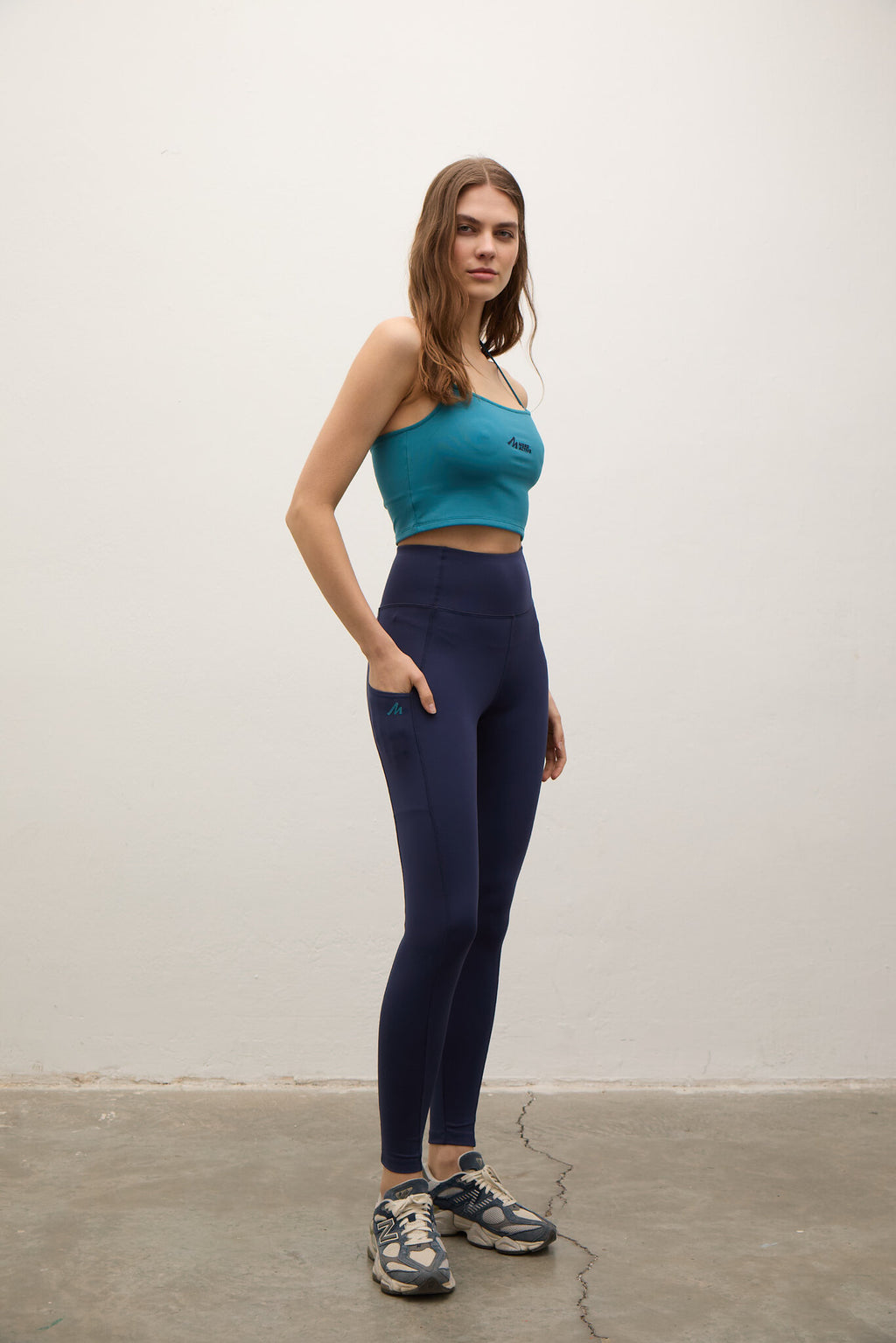 A woman stands in a studio wearing a teal sports bra and navy blue high-waisted leggings with side pockets, paired with gray and blue athletic sneakers.