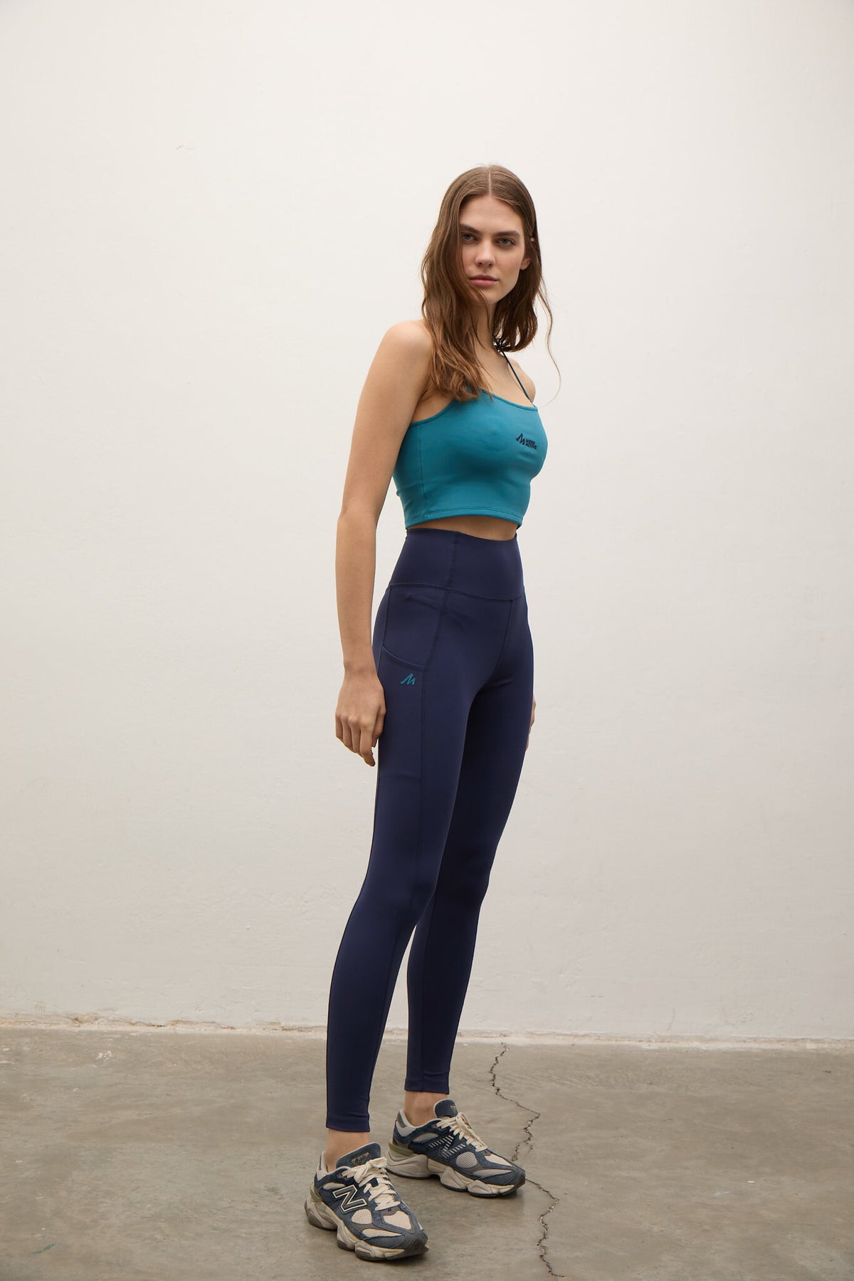 A woman modeling a matching activewear set consisting of a teal tank top and navy blue high-waisted leggings, paired with gray athletic sneakers, standing against a plain off-white wall.