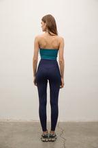 A woman stands with her back to the camera wearing high-waisted navy blue leggings and a teal strapless tube top, standing on a concrete floor against a plain off-white wall.